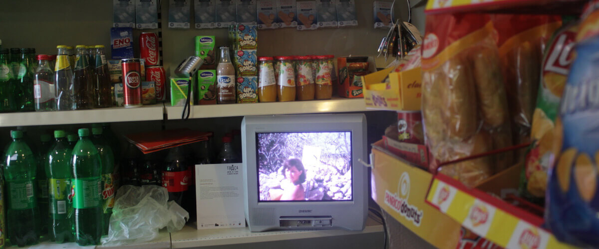 Imagem de um dos filmes do arquivo Traça. Fotografia de uma pequena televisão numa prateleira de supermercado, rodeada pelos produtos à venda, de vinho a batatas fritas de pacote.