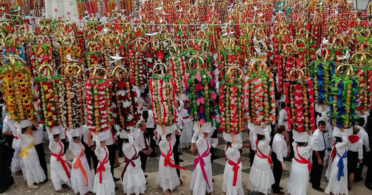 Fotografia do Cortejo dos Tabuleiros em Julho de 2019. As mulheres, vestidas de branco e com grinaldas coloridas, transportam altos tabuleiros compostos por pão e flores, encimados por uma coroa com a pomba branca, que representa o espírito santo.