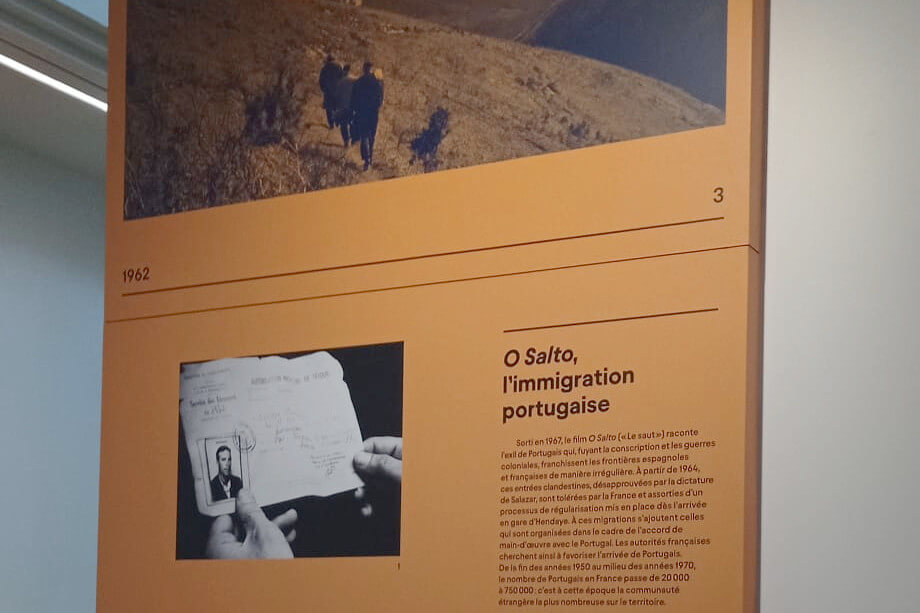 Fotografia de um dos painéis da exposição com o título “O Salto, l’immigration portugaise”. O texto do painel é ilegível, mas vêem-se duas fotos: uma de pessoas a caminhar num trilho de montanha e outra de um documento de identificação.