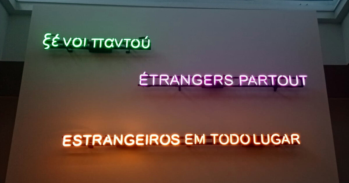 Fotografia de uma parede da exposição onde se encontram três neons com o texto “Estrangeiros em todo lugar” em português (cor laranja), francês (cor lilás) e grego (cor verde).