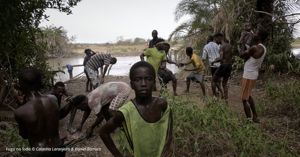 Imagem do filme "Fogo no Lodo" de Catarina Laranjeiro e Daniel Barroca, onde se vêm um grupo de meninos e homens adultos negros junto a um rio. Em primeiro plano, um menino olha directamente para a câmara.