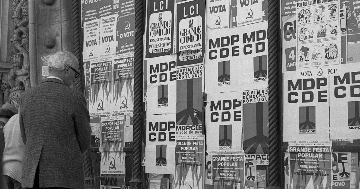 Detalhe de uma fotografia, da autoria de Diogo Margarido, onde se vê um homem de cabelos brancos em frente a uma parede, no exterior da estação do Rossio, repleta de cartazes de diversas organizações: Partido Comunista Português, MDP CDE, LCI. Há cartazes de apelo ao voto, outros de anúncios de comícios e outros de festas populares.