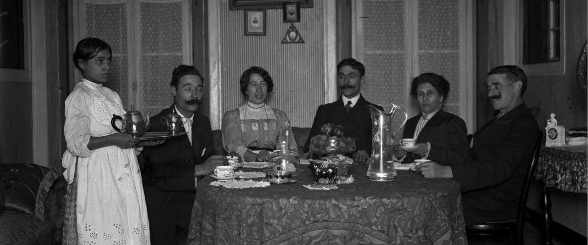fotografia que mostra uma família (três homens com bigode e duas mulheres) sentada à mesa, de frente para a câmara, com uma jovem rapariga, de pé, a segurar uma bandeja com um bule de chá; na mesa, estão distribuídas chávenas e jarros. A foto é da autoria de Eduardo Alexandre da Cunha e será do início do século 20.