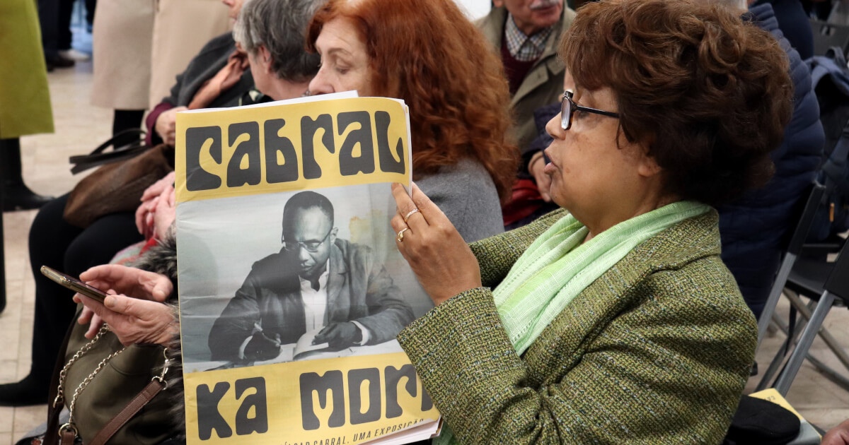 Fotografia de uma mulher a ler o catálogo-jornal de “Amílcar Cabral, Uma exposição”, com a capa bem visível, onde se pode ler o título “Cabral Ka Mori” e ver uma foto de Amilcar Cabral, sentado a escrever. A mulher fotografada encontra-se sentada entre o público que assistia ao debate de inauguração da exposição em Lisboa.