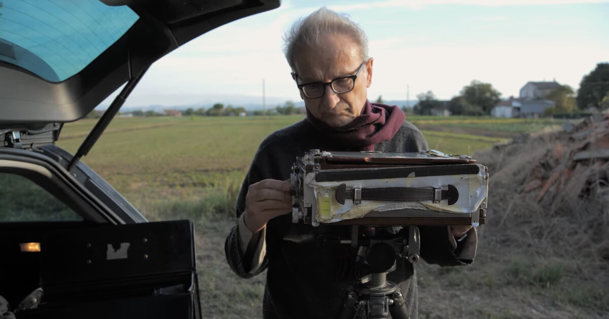 Fotograma do filme “Vivi Nascosto Guido Guidi” que mostra Guido Guidi, junto a uma mala de carro aberta, a montar uma câmara fotográfica de fole.