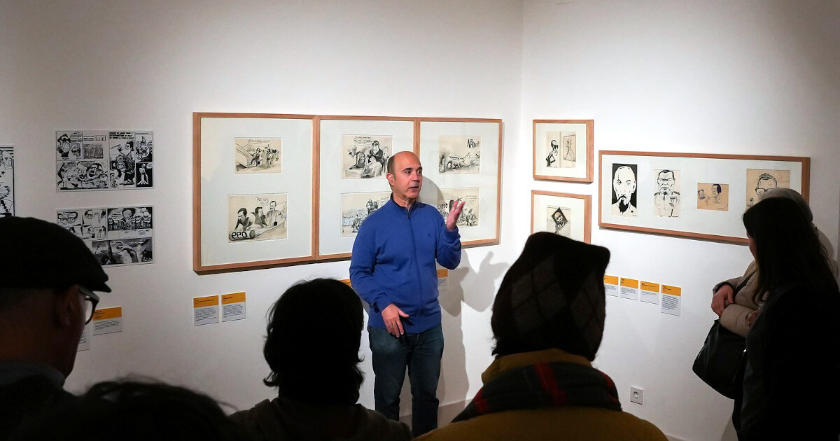 Fotografia de Paulo Jorge Fernandes a fazer a visita inaugural à exposição numa sala com muitos cartoons emoldurados nas paredes.