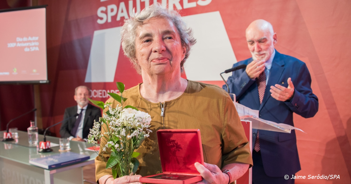 Fotografia da Irene Flunser Pimentel na cerimónia de homenagem com um ramo de flores brancas na mão e uma caixa aberta com a Medalha de Honra da Sociedade Portuguesa de Autores. Atrás da Irene, ainda no púlpito, está José Jorge Leiria, presidente da Sociedade Portuguesa de Autores.