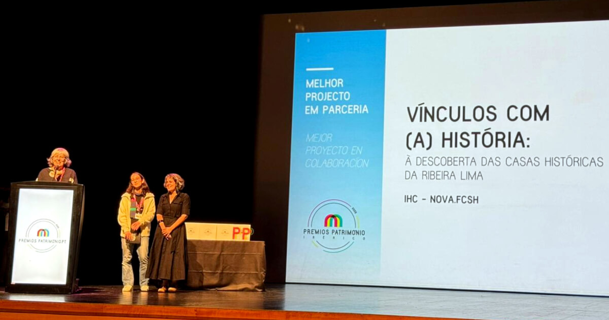 Maria de Lurdes Rosa, acompanhada de Rita Sampaio da Nóvoa e da Mafalda, no palco dos Prémios Património Ibérico 2025. Atrás delas, no ecrã, está projectada uma imagem com os textos: “Melhor projecto em parceria”, “Vínculos com (a) História: à descoberta das casas históricas da Ribeira Lima” e IHC — Nova FCSH.